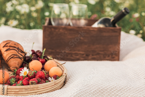 a tray with croissants, strawberries, cherries and apricots in the background is a box with a bottle of wine and two glasses. Picnic in the chamomile field