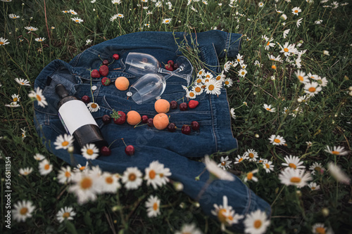 a bottle of wine, two glasses, cherries, strawberries on a denim jacket in a chamomile field
