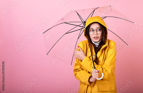 Sexual student in a yellow cloak with a hood on her head with glasses under an umbrella, snuggles her hands because of the cold on a pink background.