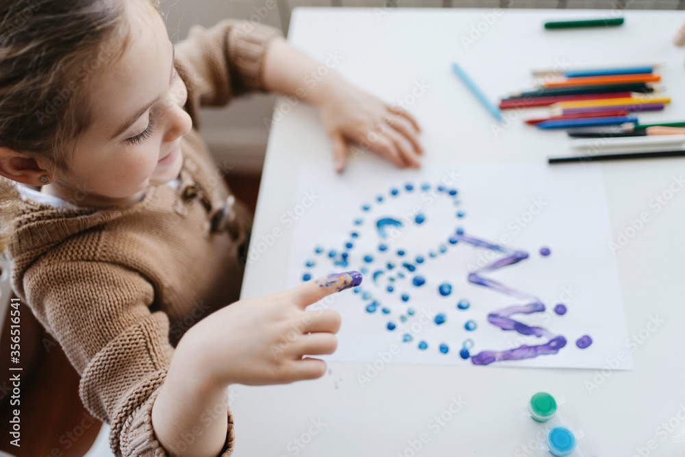 Cute child in casual outfit drawing paints with fingers while sitting at table with colour pencils and scissors at light kindergarten Cute child in casual outfit drawing paints with fingers while sitting at table with colour pencils and scissors at light kindergarten