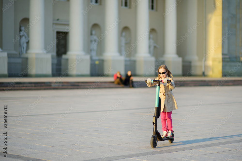 Cute young girl riding an electric scooter on sunny spring day. Electric urban transportation in Vilnius, Lithuania. Scooters for rent. Family leisure with kids.