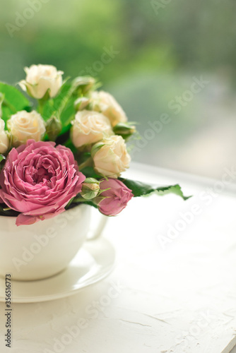 A bouquet of white roses in a cup on a white table opposite the window.