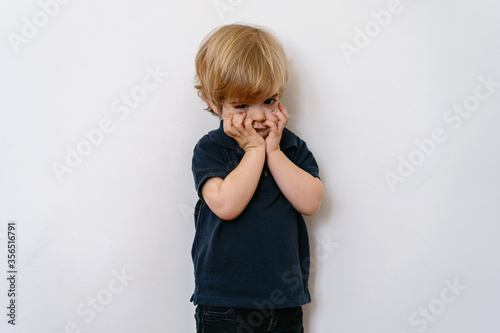 Annoyed blond little boy in casual clothes looking away with dissatisfaction while standing on white wall with hands on face photo