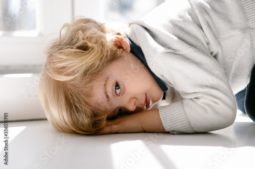 Side view of calm blond boy in casual wear getting looking at camera while resting on windowsill in modern apartment on sunny day photo