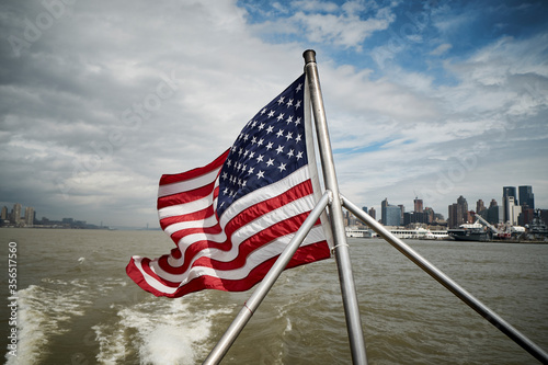 National flag of USA waving on pole of floating vessel against cloudy sky near New York City coast