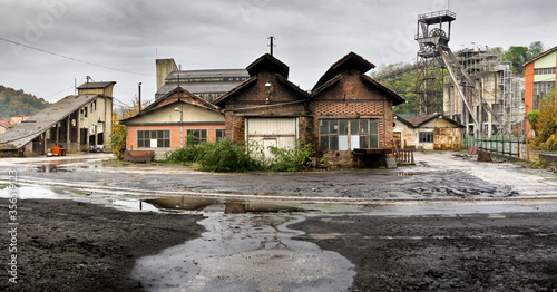 Wet uneven asphalt and shabby old one story deserted wooden houses on background of green trees and abandoned coal mine on cloudy cool day after rain