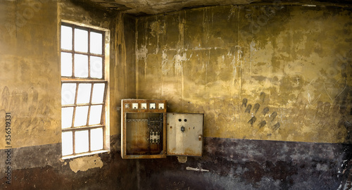 Old switch box installed in corner of weathered room with shabby stone walls and window with grid inside desolated industrial building