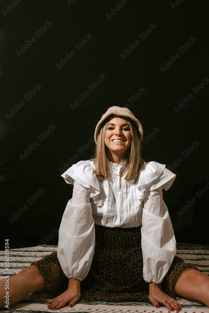 Cheerful young cute blonde female in stylish outfit and hat sitting on floor and smiling for camera against black background Cheerful young cute blonde female in stylish outfit and hat sitting on floor and smiling for camera against black background