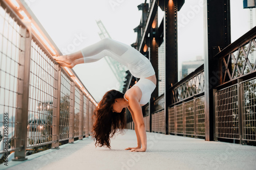 Low angle side view of flexible female in sports bra and leggings performing scorpion handstand while exercising barefoot and leaning on metal railing in city