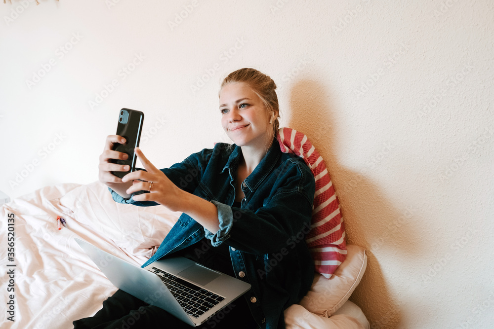 Young happy female in wireless earphones and denim jacket using smartphone to take a selfie and laptop while relaxing on bed in modern apartment Young happy female in wireless earphones and denim jacket using smartphone to take a selfie and laptop while relaxing on bed in modern apartment