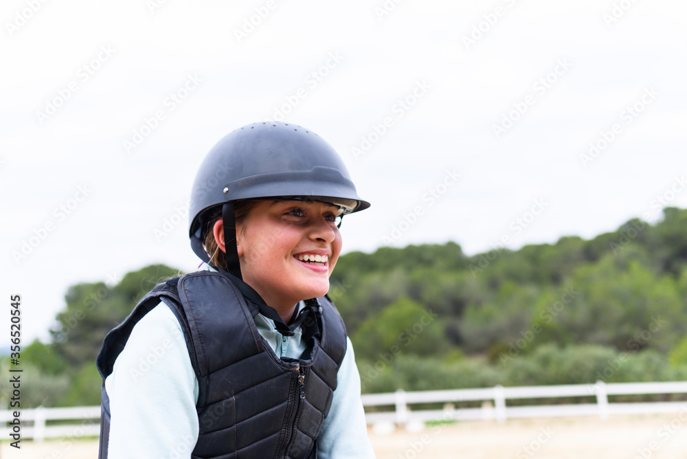 Cheerful teen girl in jockey helmet and waistcoat smiling and looking away during horseback riding lesson in equestrian school in countryside Cheerful teen girl in jockey helmet and waistcoat smiling and looking away during horseback riding lesson in equestrian school in countryside