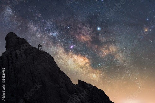 From below of silhouette of traveler standing on rough rock and looking through telescope while enjoying stunning view of amazing starry sky with breaking rays of rising sun