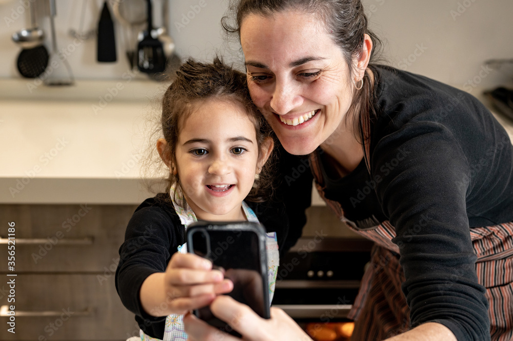 Happy woman in apron smiling and taking selfie with mobile phone with happy little girl while cooking pastry together in cozy kitchen at home Happy woman in apron smiling and taking selfie with mobile phone with happy little girl while cooking pastry together in cozy kitchen at home