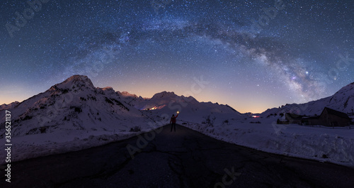 Breathtaking view of distant silhouette of explorer standing on road leading to snowy mountainous area and holding luminous light in hand against starry stunningly beautiful sky on winter night photo