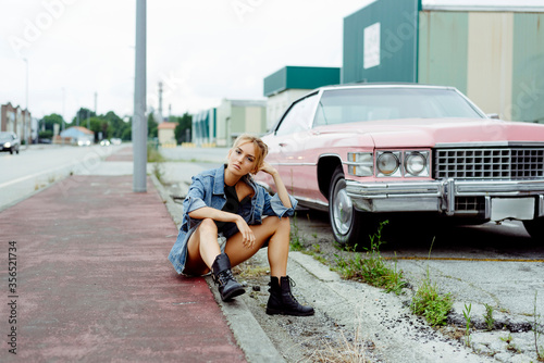 Attractive blonde girl sitting on the sidewalk near to a classic pink car