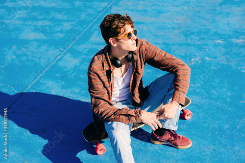 From above confident young man in casual clothes sitting on skateboard and looking away on sunny day in skate park photo