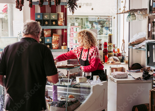 Senior man choosing food from freezer while buying wine and cheese from female seller in cozy small delicatessen store