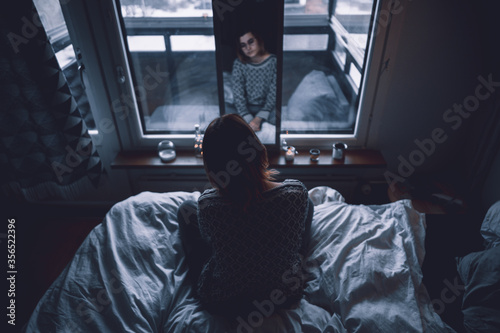 Back view of depressed young female sitting on bed in dark bedroom looking at mirror at home