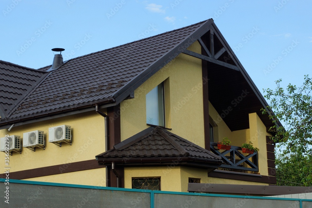 part of a yellow private house with windows and a wooden open balcony under a brown tiled roof against a blue sky part of a yellow private house with windows and a wooden open balcony under a brown tiled roof against a blue sky