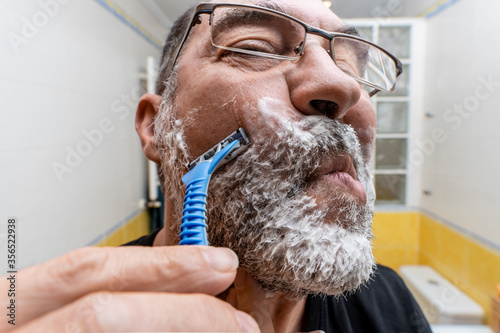 Low angle closeup of middle aged bearded male shaving with disposable razor in front of mirror in bathroom