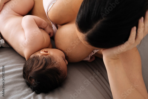 Peaceful female in white underwear lying down on comfortable bed and breastfeeding little cute baby