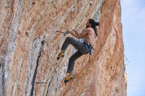 From below side view of shirtless bearded alpinist in safety equipment climbing up on rocky slope of mountain and looking up