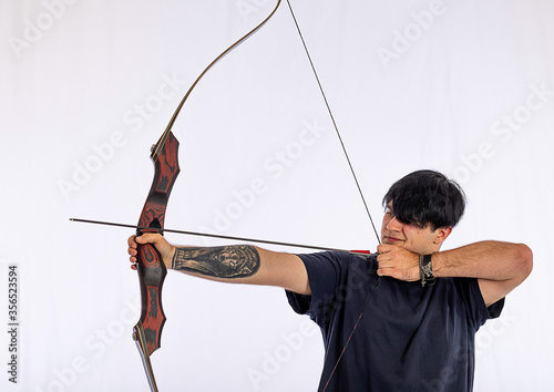 Portrait of a European young man, 21 years old with wolfs tattoo on arm, against a white background, spans a takedown recurved bow and aims