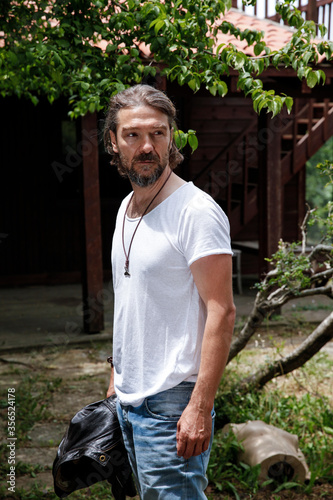 Handsome man in his 40's posing in a garden. Bearded man holding a black leather jacket in his hand and looking away with a serious face. Wearing white T-shirt and denim jeans.