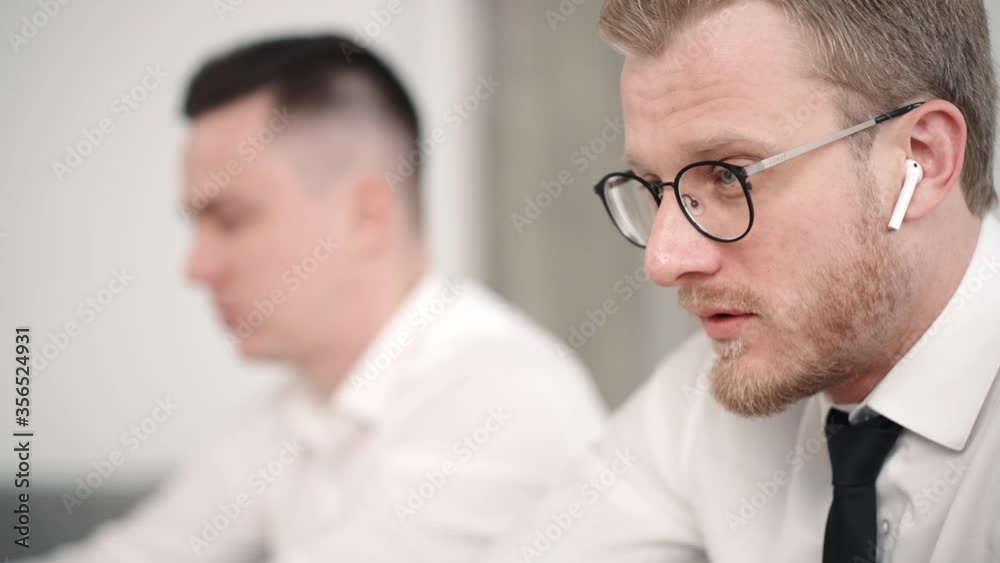 two modern business men working together in office look at laptop screen and talk. wear wireless headphone. conversation between colleagues. two coworkers discuss project two modern business men working together in office look at laptop screen and talk. wear wireless headphone. conversation between colleagues. two coworkers discuss project