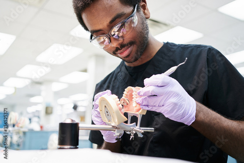 African American guy in latex gloves using mouth mirror and probe to check false teeth while working in modern laboratory photo