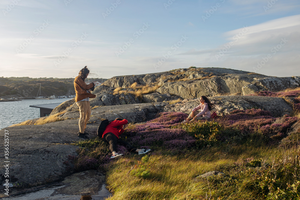 Group of tourist in casual clothes standing on stone and taking pictures of friend on photo camera on rocky seashore Group of tourist in casual clothes standing on stone and taking pictures of friend on photo camera on rocky seashore