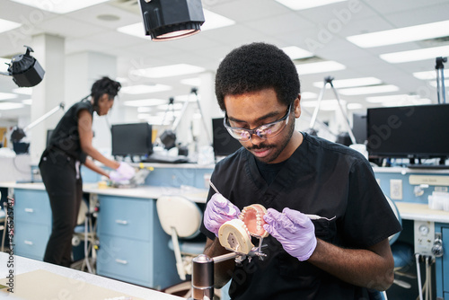 African American guy in latex gloves using mouth mirror and probe to check false teeth while working in modern laboratory