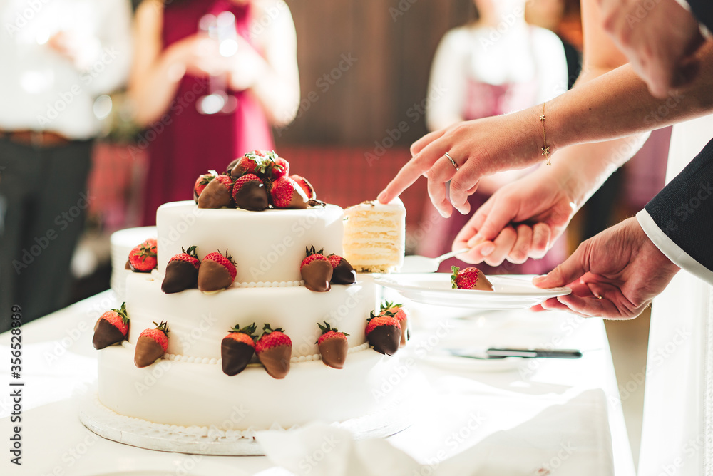 Bride slicing white wedding cake decorated with strawberries and serving one piece on white plate held by groom. Newlyweds cooperation. Wedding day
