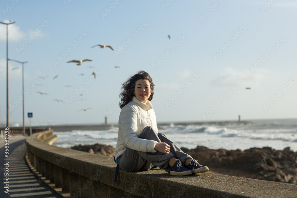 Mixed race woman sitting of sea promenade in Porto, Portugal. Seagulls fly over the foaming surf.