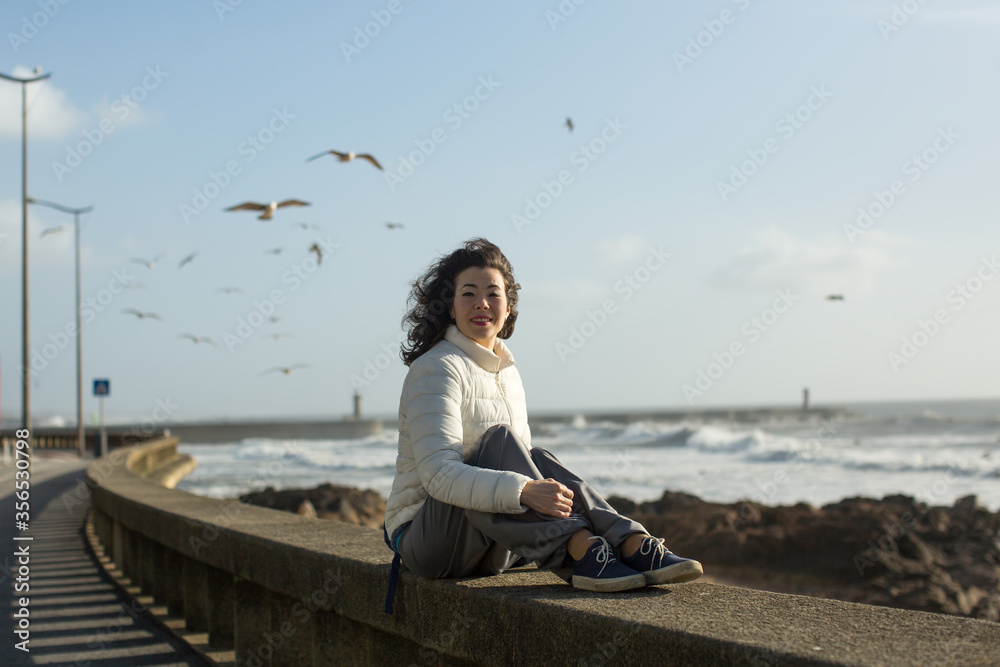 Mixed race woman sitting of sea promenade in Porto, Portugal. Seagulls fly over the foaming surf. Mixed race woman sitting of sea promenade in Porto, Portugal. Seagulls fly over the foaming surf.