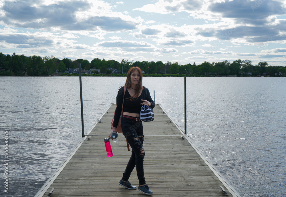 Happy red-haired girl dressed in black with a striped bag, a pink backpack and two bottles of water in her hand standing in the middle of a pier in front of a large lake ready for a picnic