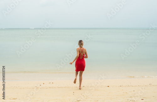 Young woman in a red dress runs and jumps on a white sand beach into tropical sea. Young happy woman running on the beach. Happiness to be on a sea or ocean. Vacation and travel concept.