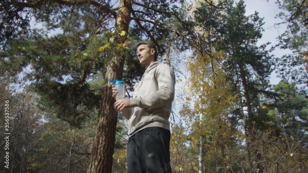 Healthy sportsman rests between exercises in gorgeous autumn forest drinking with delight from training bottle. Low angle 4K 360 degree tracking arc shot. Healthy sportsman rests between exercises in gorgeous autumn forest drinking with delight from training bottle. Low angle 4K 360 degree tracking arc shot.
