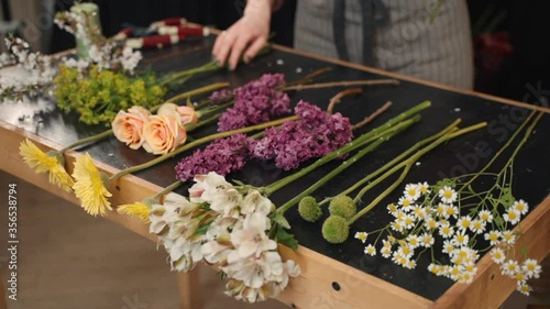 Young woman takes flowers in her hands to create composition while standing in workshop. Female florist is working with fresh blossoms lying on table in light interior of store. Creative professional