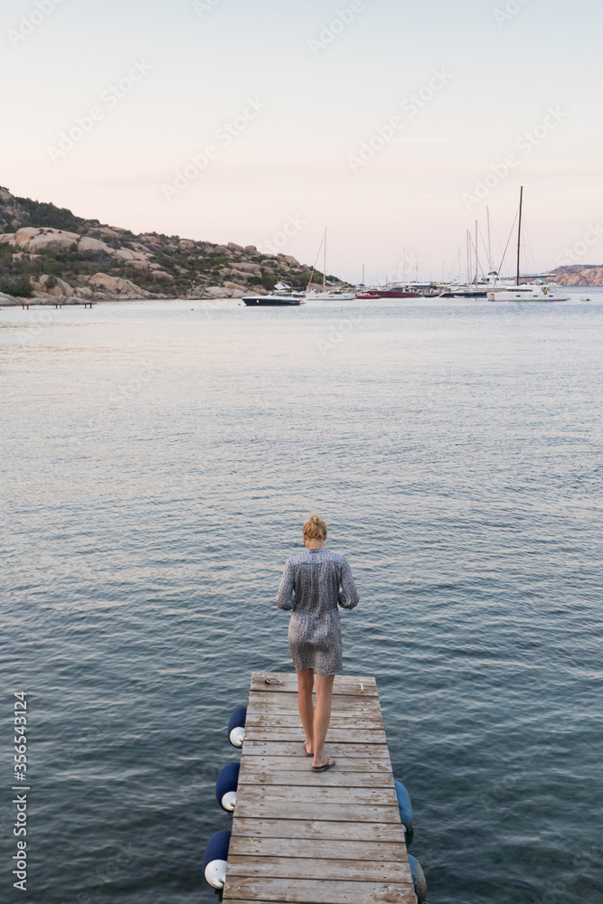 Beautiful woman in luxury summer dress standing on wooden pier enjoying peaceful seascape at dusk. Female traveler stands on a wooden pier in Porto Rafael, Costa Smeralda, Sardinia, Italy.