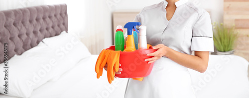 Young chambermaid holding cleaning supplies in bedroom, closeup view with space for text. Banner design