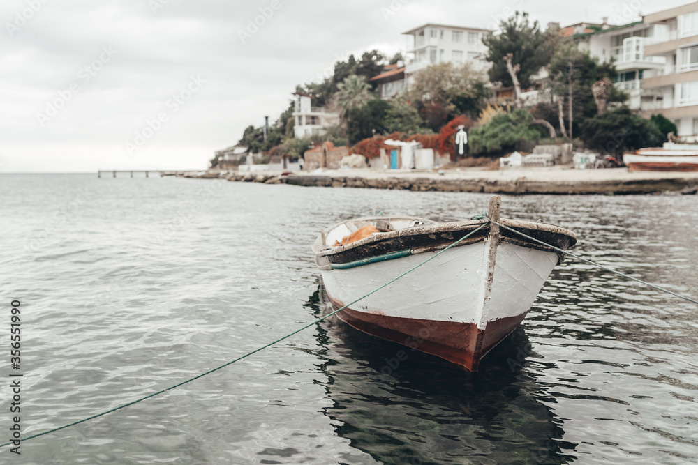An old white wooden boat by the sea is moored in a quiet bay against a background of white houses, green trees, gray cloudy sky and rocky shore. beautiful sea view with ship, pier and city in Istanbul