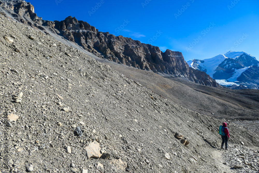 A woman trekking to the top of Thorung La Pass, Annapurna Circuit Trek, Nepal. Harsh and barren landscape around. Clear and blue sky. Snow capped mountains. Early morning and cold temperature.