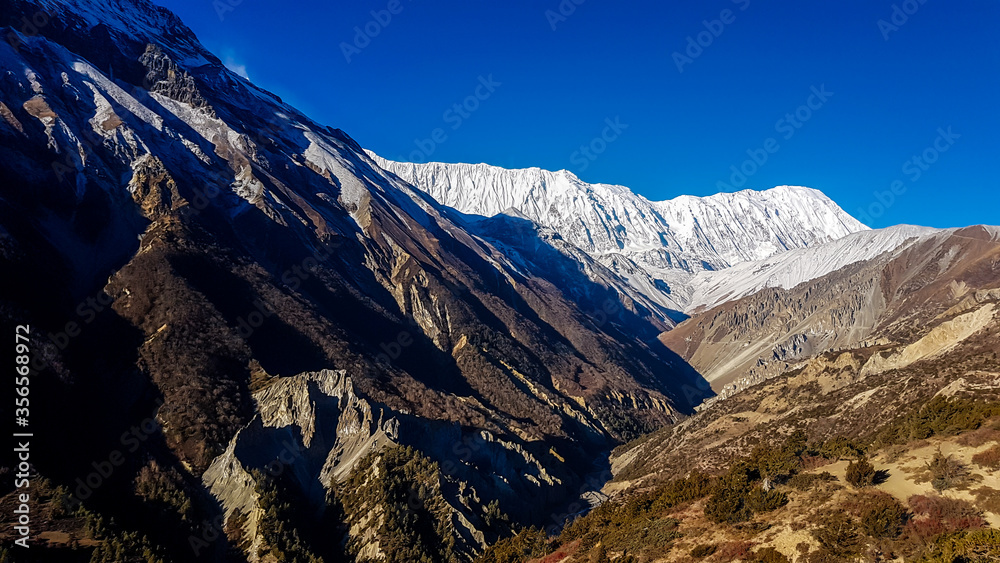 Harsh and golden colored slopes in Manang Valley, Annapurna Circus Trek, Himalayas, Nepal, with the view on Annapurna Chain and Gangapurna. Dry and desolated landscape. High snow capped mountain peaks