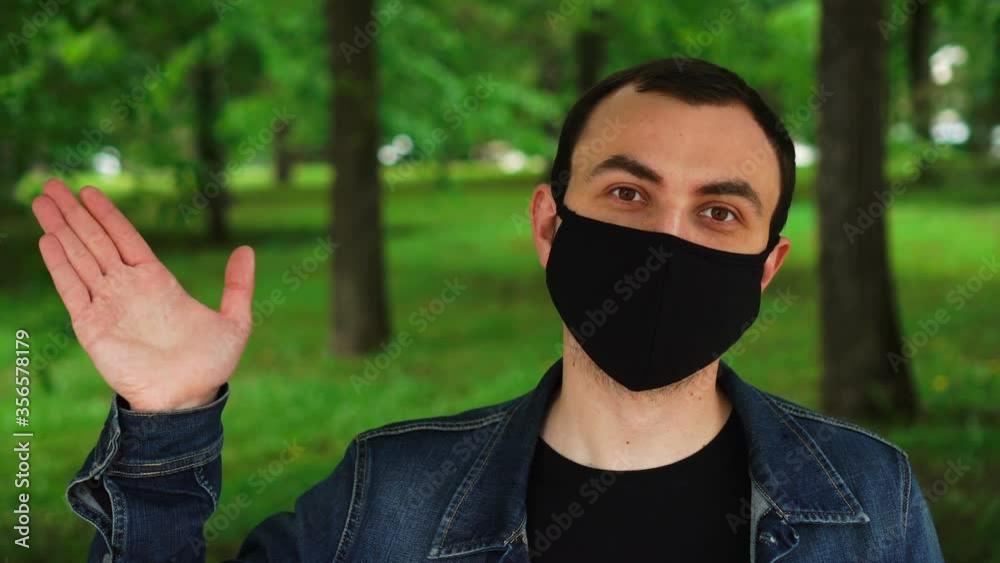 Handsome young man standing outdoors wearing medical mask and saluting by waving his hand Handsome young man standing outdoors wearing medical mask and saluting by waving his hand