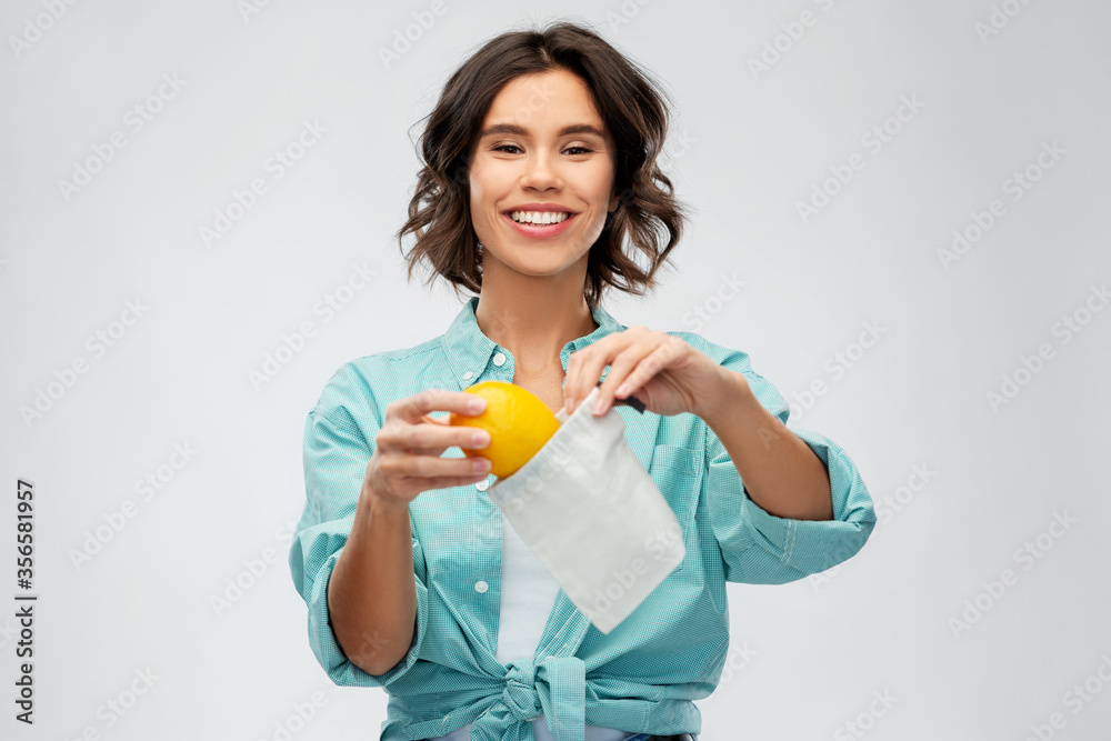 sustainability, eco living and people concept - portrait of happy smiling young woman in turquoise shirt putting orange into reusable bag over grey background sustainability, eco living and people concept - portrait of happy smiling young woman in turquoise shirt putting orange into reusable bag over grey background