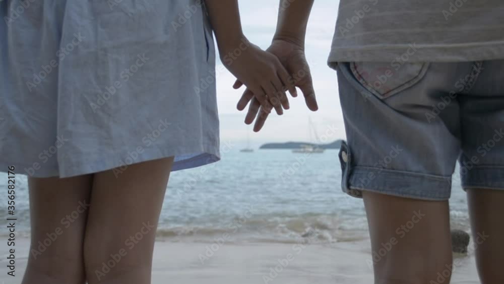 Mother and her daughter holding hands together while standing on the beach. View from rear side of female teenager and her mom watching beautiful scenery of the sea in the evening. Tilting shot. Mother and her daughter holding hands together while standing on the beach. View from rear side of female teenager and her mom watching beautiful scenery of the sea in the evening. Tilting shot.