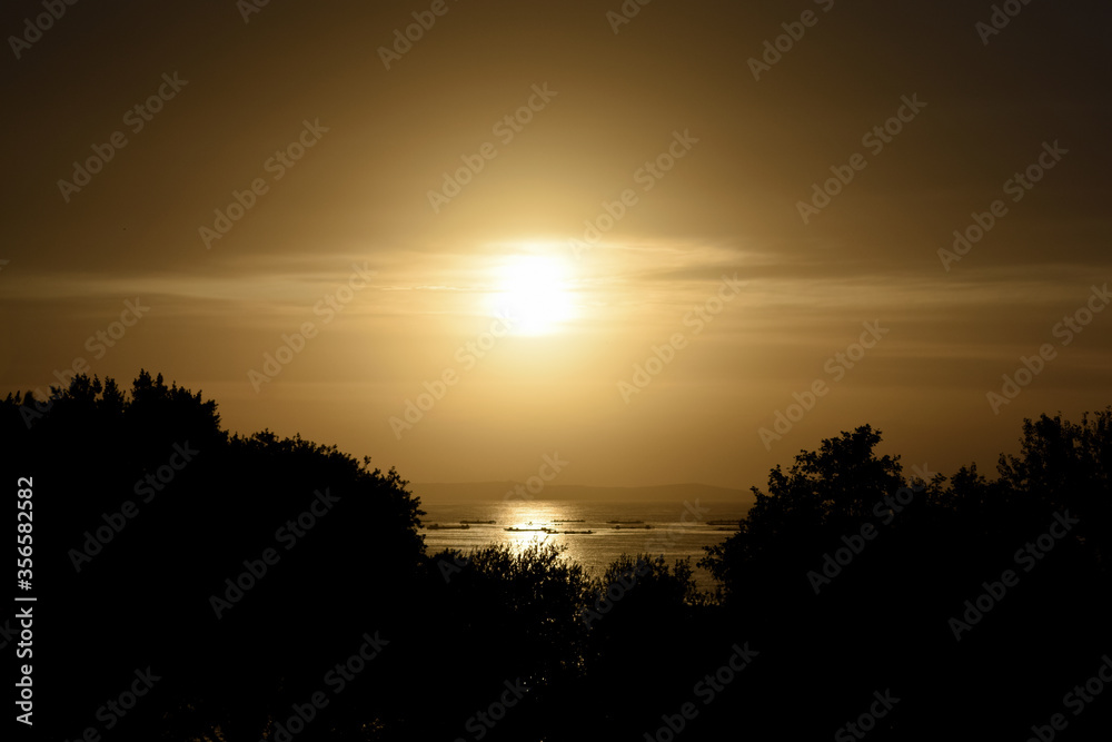 Cálida y dorada puesta de sol sobre el bosque y el horizonte de la isla de Ons (Ría de Pontevedra - España). Siluetas de mejilloneras gallegas sobre el mar brillante y dorado.