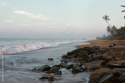 Beautiful postcard landscape shot of the scenic Pitiwella beach on sunrise with orange sand, green palm trees, rocks, the Indian ocean with big white foamy waves and the shore on the horizon. photo