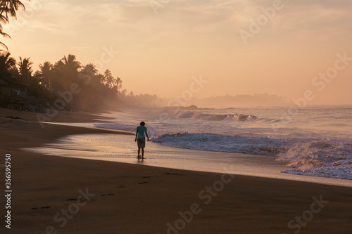 Beautiful postcard landscape shot of a woman walking on Pitiwella beach (Sri Lanka) at sunrise with orange sand, green palm trees, rocks, Indian ocean with white waves and the sun on the horizon